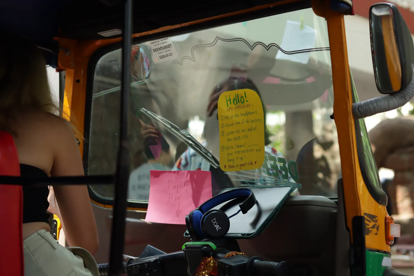 The auto's windscreen seen from inside, a yellow “Hello!” note and pink slips stuck to the glass, headphones resting on the dash.