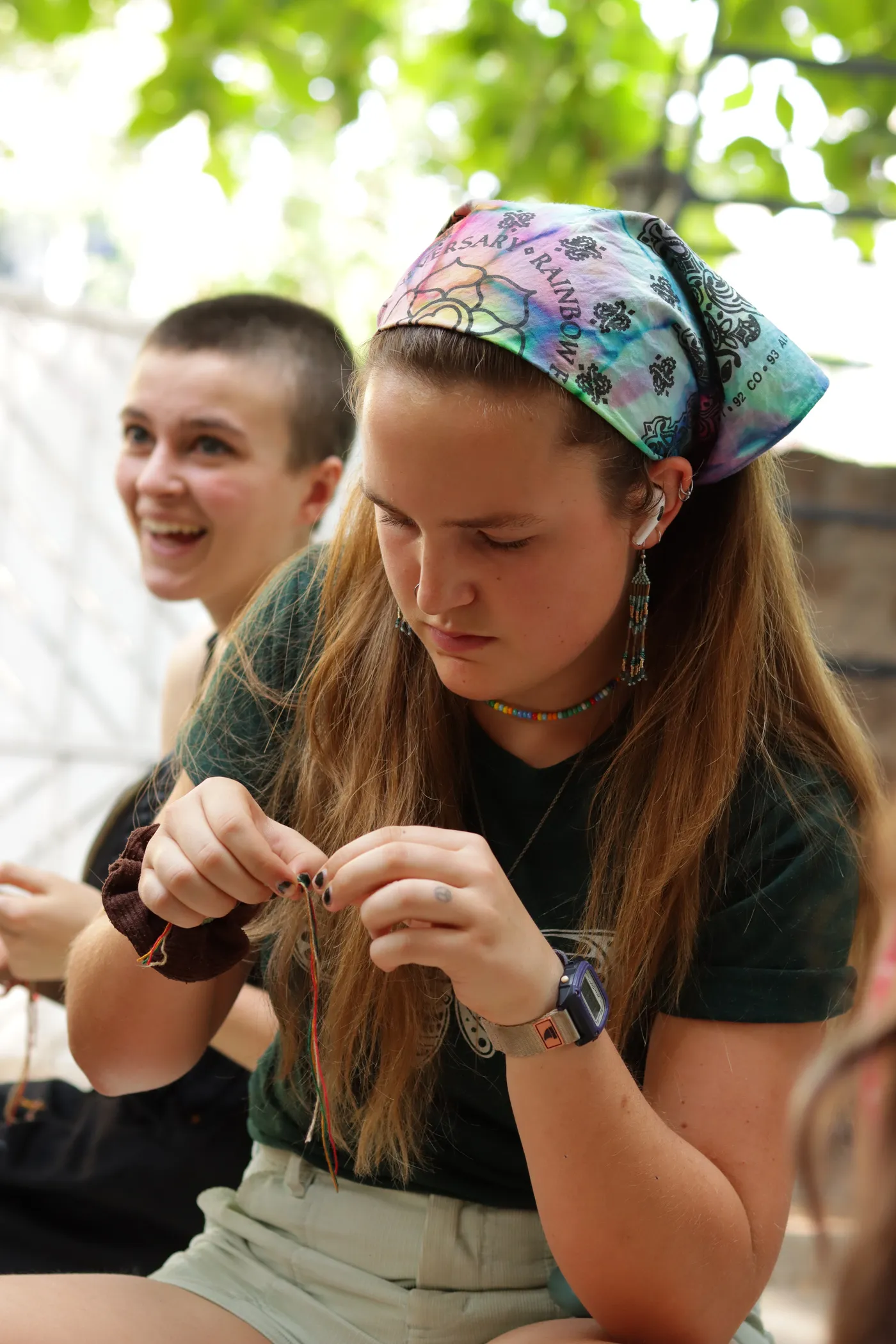 Two visitors side by side, one laughing while the other concentrates on knotting a bracelet.