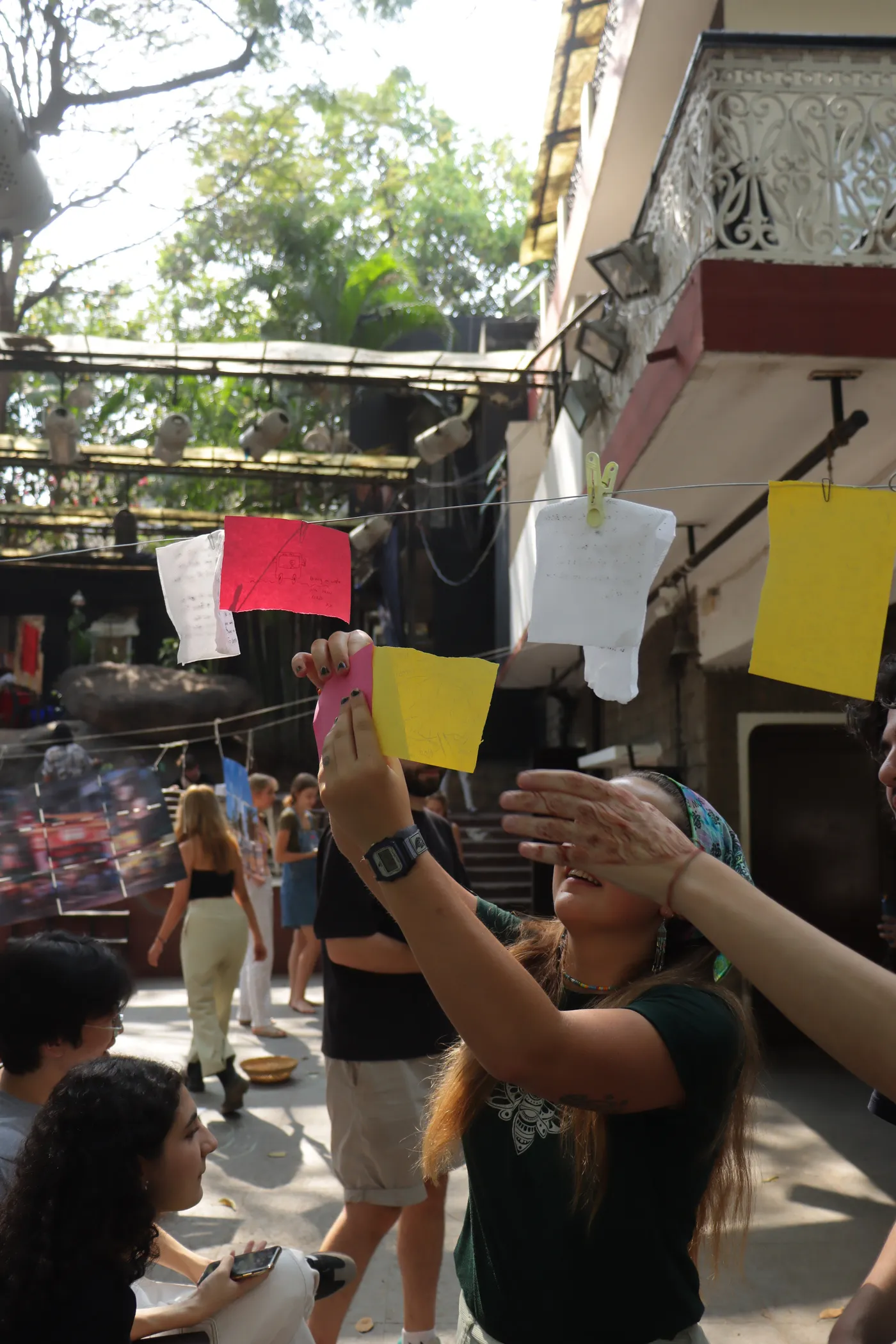 A hand reaching up to peg pink and yellow notes onto a line strung across the courtyard.