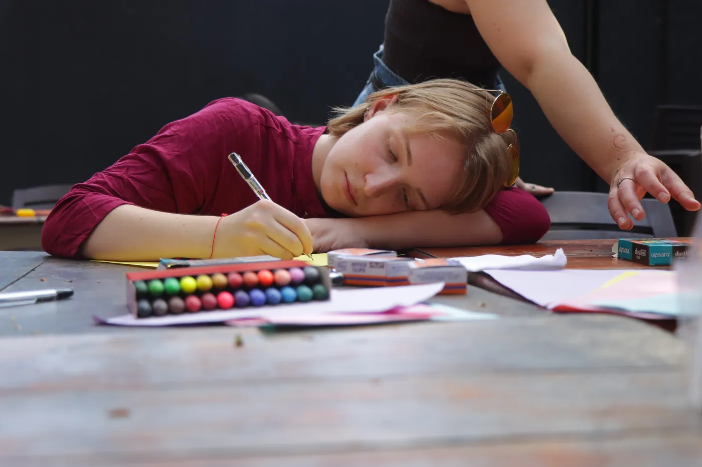 A visitor resting their head on the table as they write, a tray of coloured markers beside their hand.