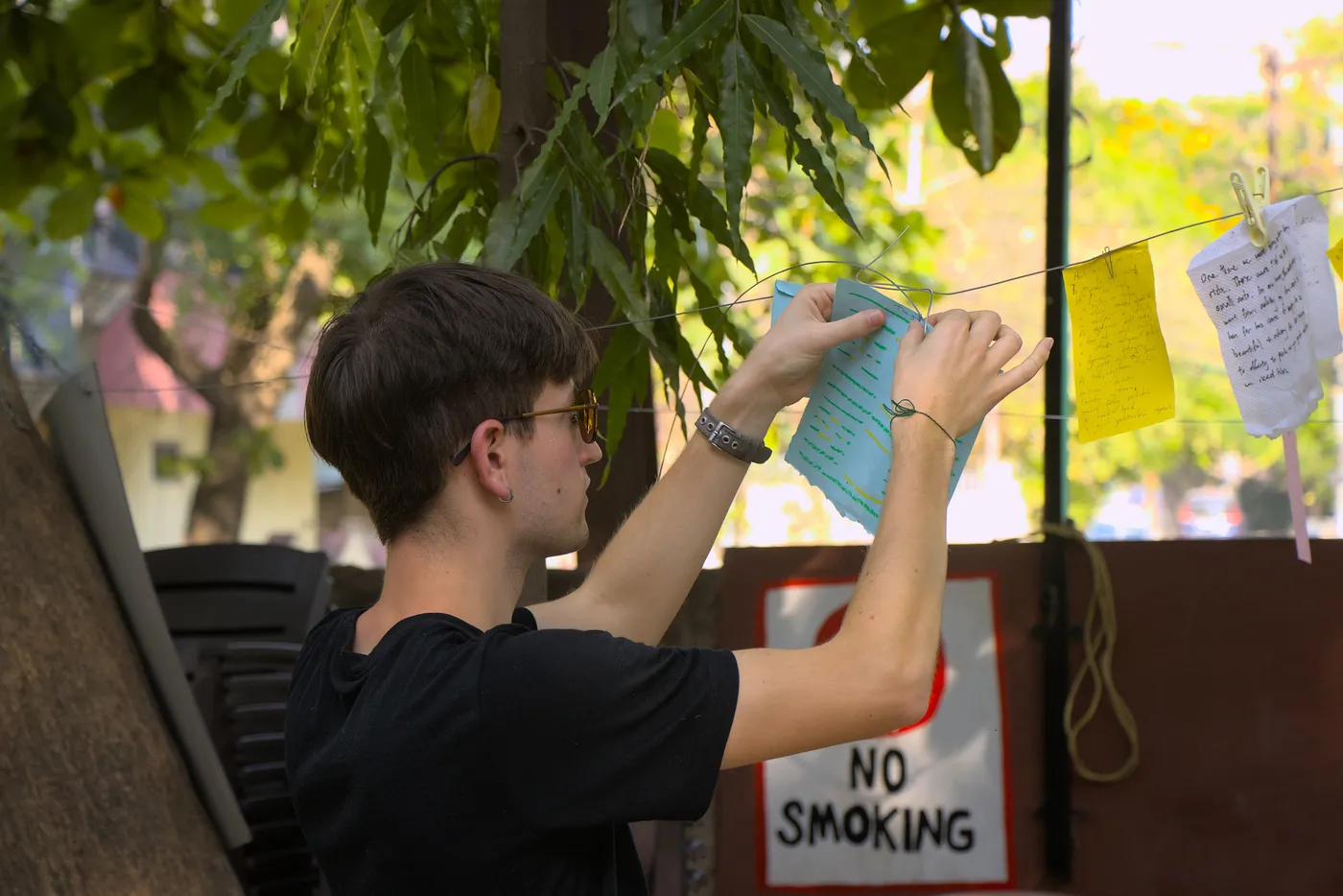 A visitor pegging a green note to a line strung under a tree, a “NO SMOKING” sign on the wall behind them.