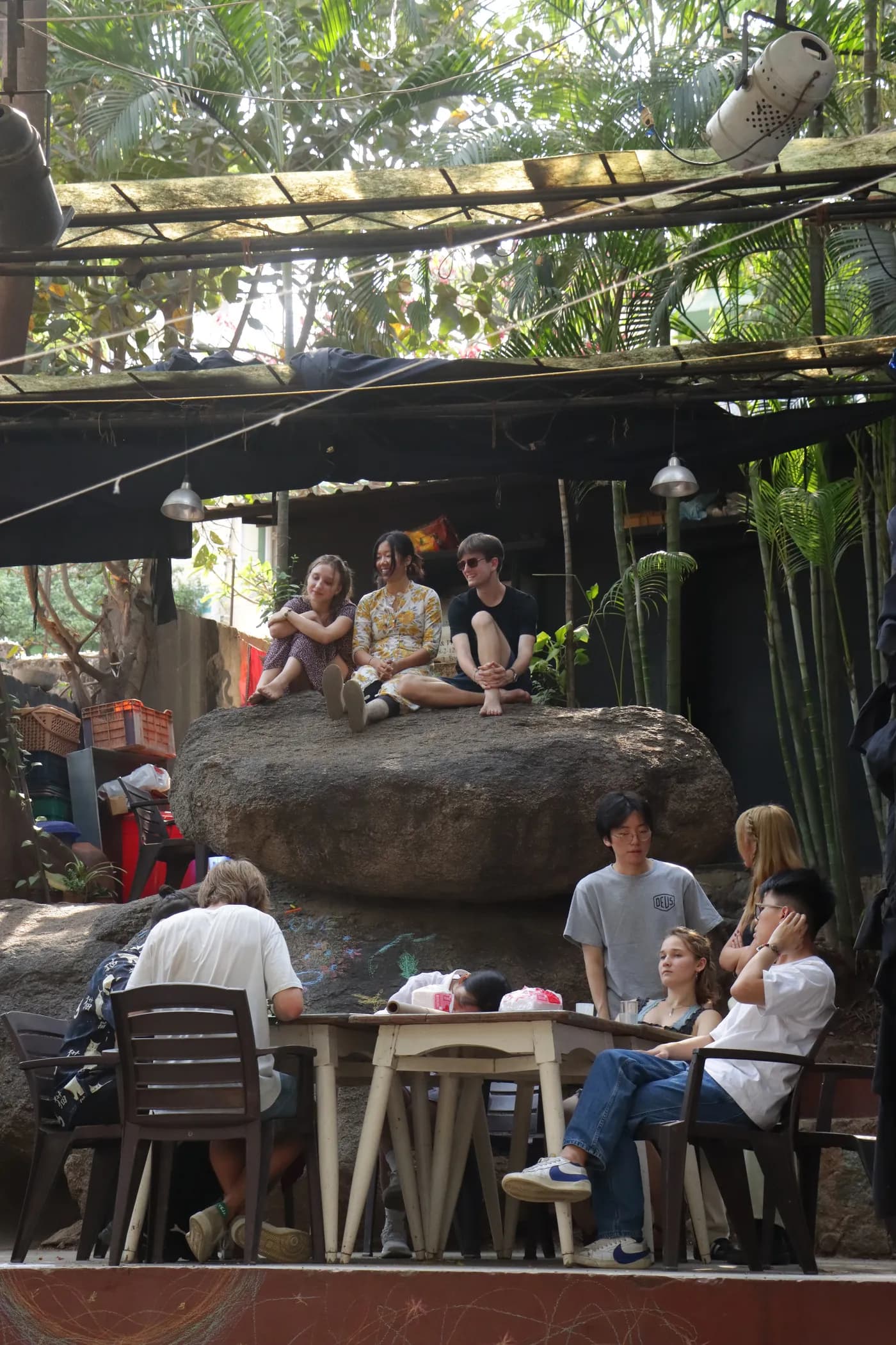 Visitors sitting on and around a large boulder in the leafy courtyard, materials spread on chairs nearby.
