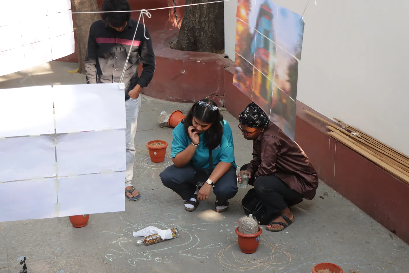 Two visitors crouched on the ground by a red wall, blank sheets pinned above them and small clay pots at their feet.