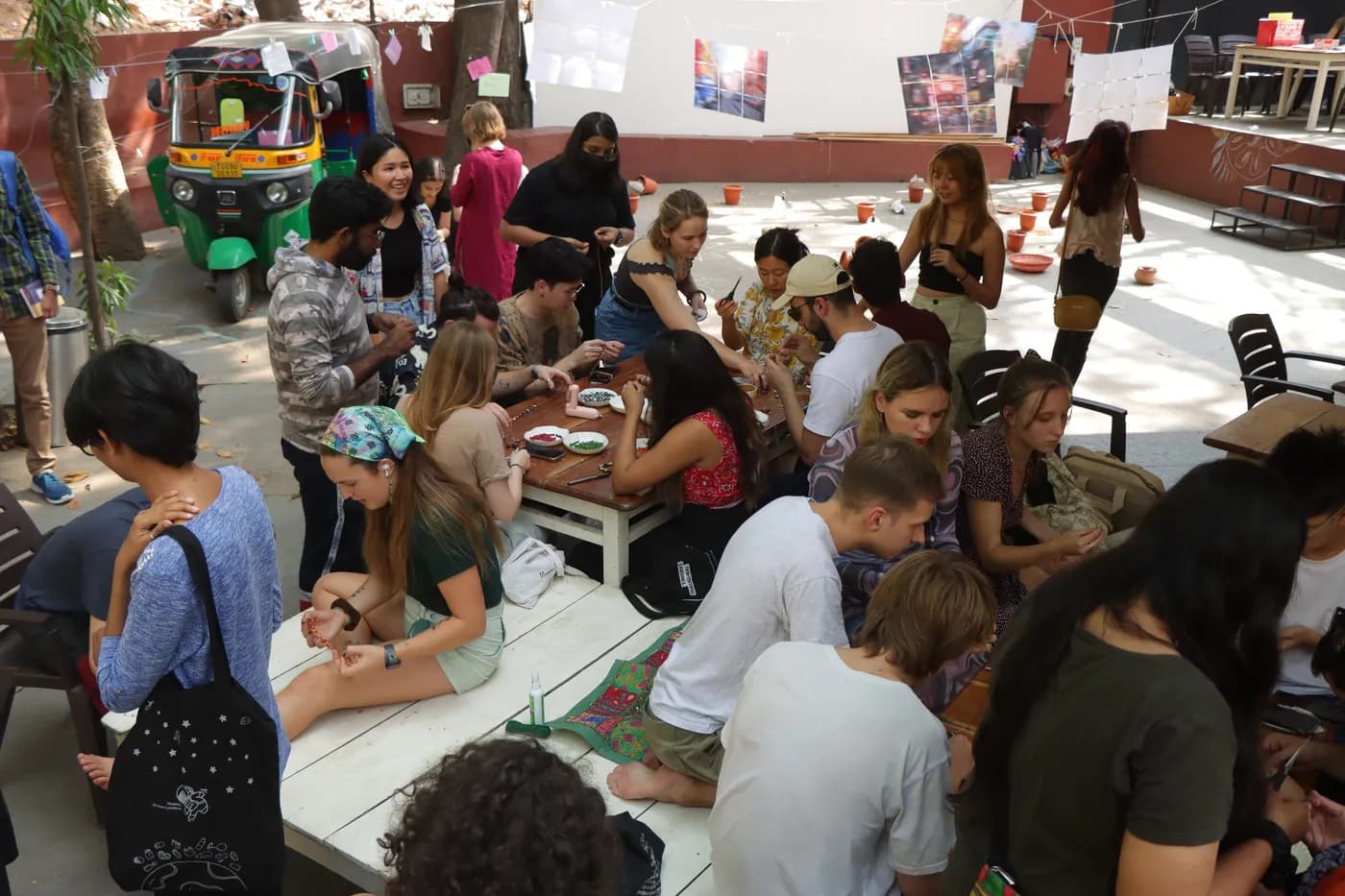 Visitors sitting close together at low tables in the courtyard, threading coloured beads onto string, with an auto-rickshaw parked behind them.