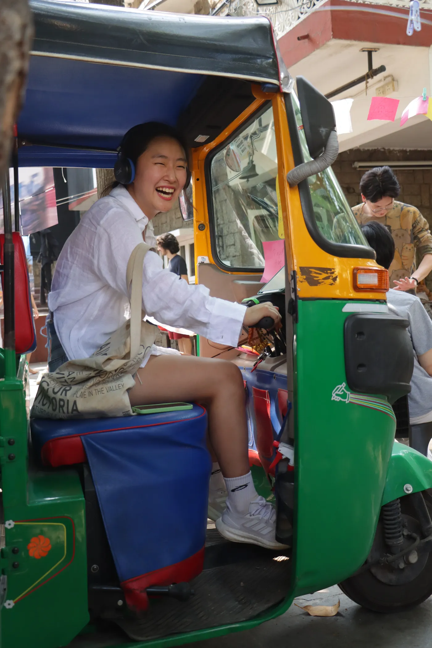 A visitor lying back across the seat of a green and yellow auto-rickshaw, laughing, with earphones in.
