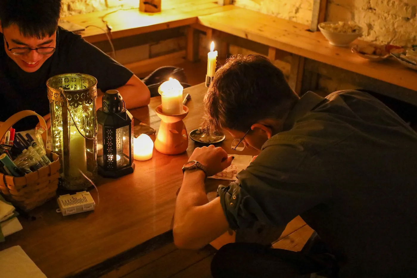 Three visitors writing at a low table by candlelight, napkins and pens spread in front of them.