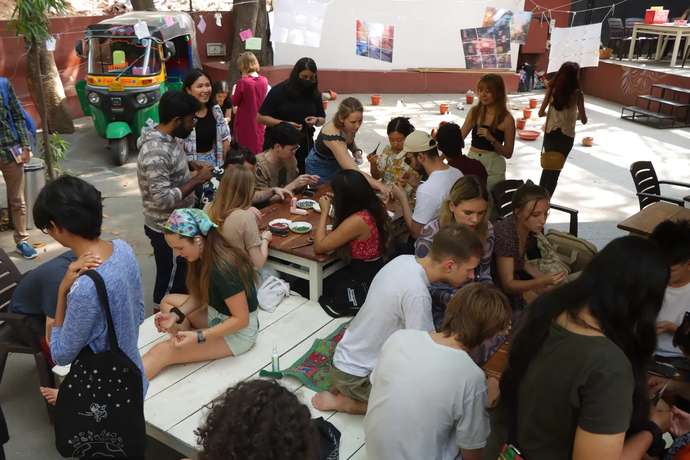 A crowd seated along low tables working on bracelets, with the autos and hanging posters behind them.